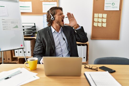 Handsome middle age man wearing call center agent headset at the office shouting and screaming loud to side with hand on mouth. communication concept.