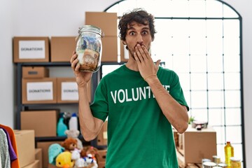 Young hispanic man wearing volunteer t shirt holding money from charity jar covering mouth with...