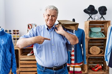Senior man with grey hair holding shopping bags at retail shop pointing finger to one self smiling happy and proud