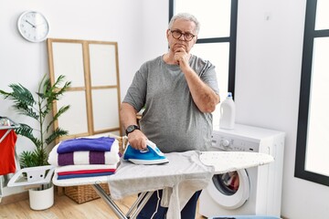 Senior caucasian man ironing clothes at home serious face thinking about question with hand on...
