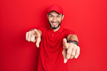 Hispanic man with beard wearing delivery uniform and cap pointing to you and the camera with...