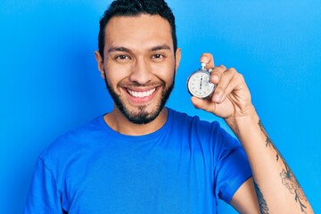 Hispanic man with beard holding stopwatch looking positive and happy standing and smiling with a confident smile showing teeth