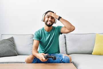 Handsome hispanic man wearing headphones playing video game holding controller stressed and frustrated with hand on head, surprised and angry face