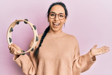 Young hispanic girl playing tambourine celebrating achievement with happy smile and winner expression with raised hand - obrazy, fototapety, plakaty