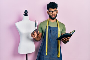 Arab man with beard dressmaker designer holding scissors and sewing kit in shock face, looking...