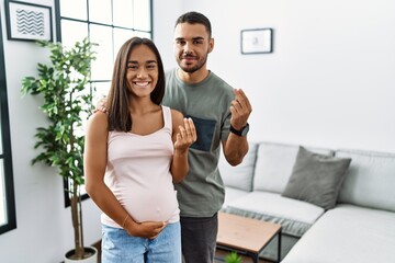 Young interracial couple expecting a baby, touching pregnant belly doing money gesture with hands,...