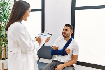 Obraz premium Youg latin injuried man wearing sling talking with the doctor at clinic waiting room.