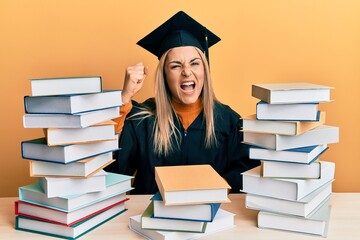 Young caucasian woman wearing graduation ceremony robe sitting on the table angry and mad raising fist frustrated and furious while shouting with anger. rage and aggressive concept.