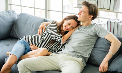 Young romantic couple embracing, sitting on cozy couch in living room relaxing at home, bonding relationship. Family portrait smiling overjoyed wife and happy closed eyes husband stay home together.