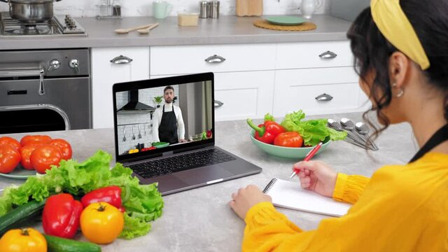 Woman in kitchen study online video call webcam laptop greets listen chef teacher, writes in notebook. Man food blogger in computer screen tells teaches housewife remote culinary lesson, slices tomato