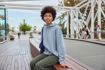 Outdoor shot of thoughtful pleased curly haired woman wears hoodie and trousers rests after...