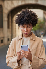 Fototapeta na wymiar Vertical shot of young curly haired woman concentrated in smartphone screen checks notification uses modern gadget for sharing media wears beige jacket poses outdoors against blurred background