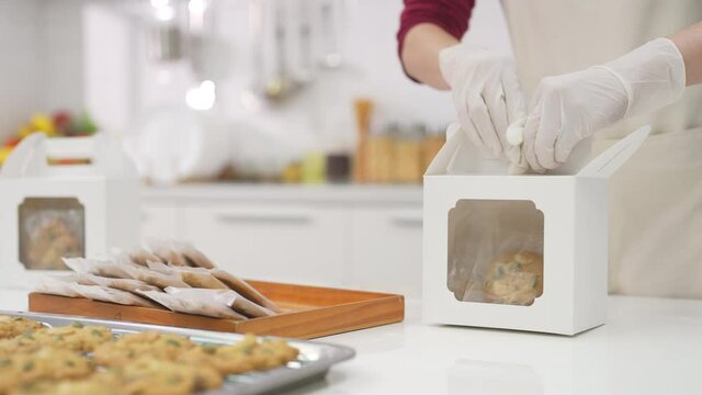 4K Asian woman pastry chef making cookie in the kitchen. Female bakery shop owner taking customer order and packing cookies in delivery box. Small business entrepreneur and food delivery concept.