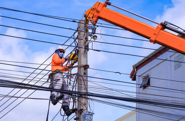 Asian electrician in protective mask with crane truck is installing cable lines and electrical transmission on electric power pole against blue sky background - obrazy, fototapety, plakaty