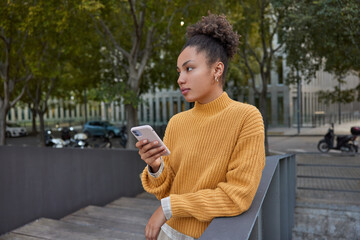 Millennial female with curly hair dressed in knitted jumper holds digital smartphone checks...