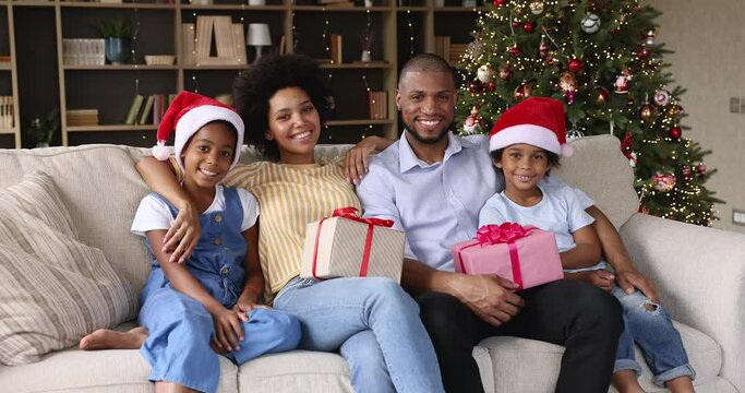 Beautiful young African couple their preschool children sit together on sofa with gift boxes near glowing Christmas tree smile look at camera. Winter holidays, Happy New Year 2022 celebration concept