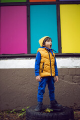 boy child in a yellow vest and hat and a blue jumpsuit stands on a tire against a background of colorful rectangles window