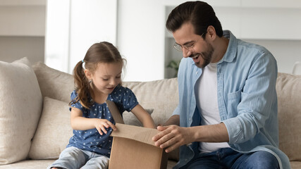 Fototapeta na wymiar Joyful adorable small child girl opening carton parcel with handsome happy young father at home, feeling excited of fast international delivery shipping, having positive online shopping experience.