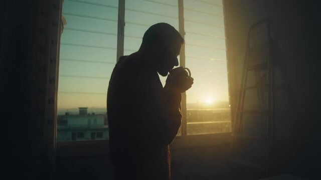 Sagoma di un uomo che beve una tazza di the alla finestra al tramonto.