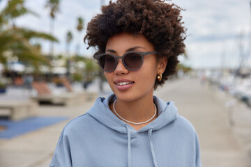Outdoor shot of pleased woman with curly hair wears sunglasses and hoodie walks at pier harbor...