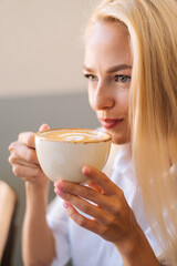 Close-up face of attractive young blonde woman drinking coffee hot cappuccino coffee with beautiful pattern sitting in cafe. Clsoeup vertical shot of cute female enjoying aroma coffee in restaurant.