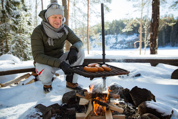 Man roasting sausages on campfire in forest by the lake, making a fire, grilling. Happy tourist...