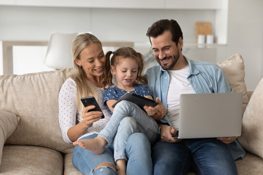 Happy bonding loving family couple parents with little preschool kid daughter holding different modern technology gadgets, spending time online sitting on comfortable couch addiction concept.
