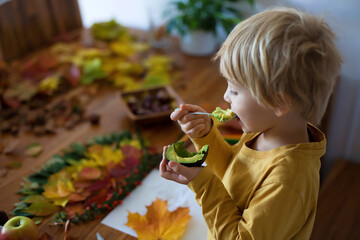 Sweet child, boy, eating avocado while applying leaves using glue while doing arts and chraft with leaves