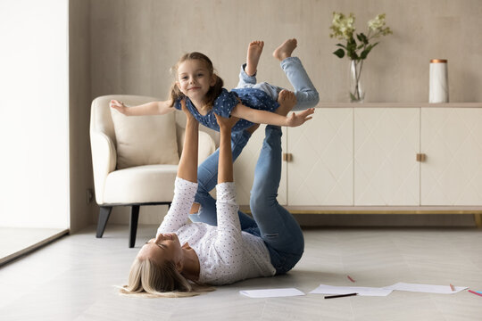 Happy young mother lifting in air laughing cute little kid daughter, lying on warm heated wooden floor in living room. Joyful two female generations family having fun, entertaining togetehr at home.