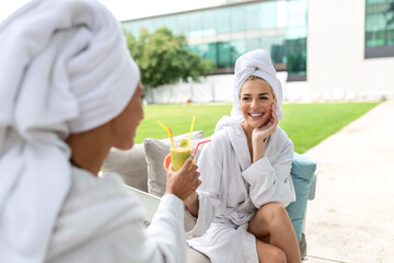 Young girlfriends relaxing in white bathrobes and drinking healthy spa cocktails outside at a...