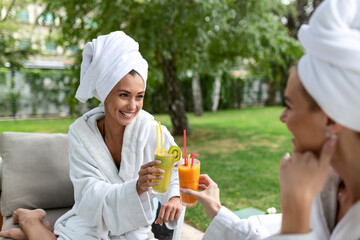 Two friendly women in white bathrobes sitting in spa with drinks and talking to each other after spa care routine