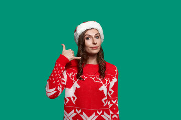 Positive european woman with dark hair encourages to call, makes phone gesture with hand, holding on the line, wears red ugly sweater with deer and santa hat on green background. Christmas concept.