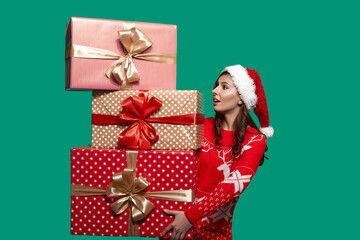 European woman with dark hair holds a pile of heavy gift boxes with bow knots, is worrying to drop it down, wears red ugly sweater with deer and santa hat on green background. Christmas concept.