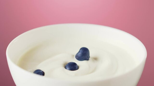 Blueberries fall into a bowl of Greek yogurt on pink background.