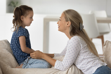 Side view smiling young woman holding hands of little cute kid daughter, involved in sincere conversation at home, helping solving problems or giving advices, trustful family relations concept.
