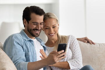 Happy bonding loving married family couple using cellphone, resting on cozy sofa at home. Affectionate young man in eyeglasses cuddling smiling wife, web surfing information or playing games.