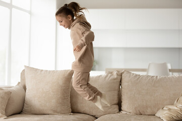 Full length joyful little 7s kid girl in comfortable loungewear jumping on sofa, having fun...