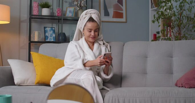 Close up of smilling woman in bathrobe, towel sitting on sofa in living room texting, chatting, reading and answering messages using smartphone, speaking to herself. Makeup kits table in living room.