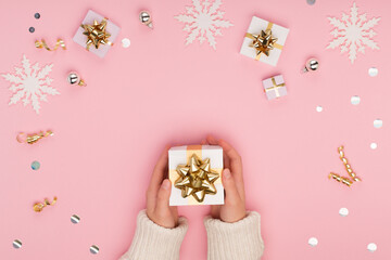 Top view on female hands holding white gift box wrapped with gold bow on Christmas pink background.
