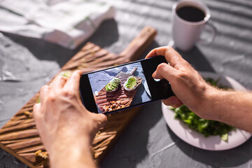 Fototapeta na wymiar Hands take pictures on smartphone of two beautiful healthy sour cream and avocado sandwiches lying on board on the table. Social media and food concept
