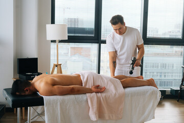 Wide shot of professional male masseur massaging leg calf muscles using massage gun percussion tool...