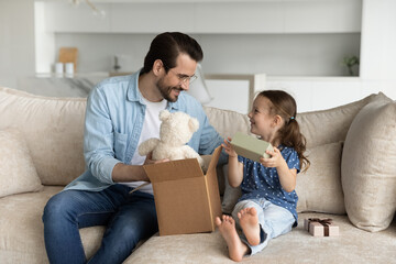 Happy young father unboxing parcel with gifts for small cute kid daughter, sitting together on cozy...
