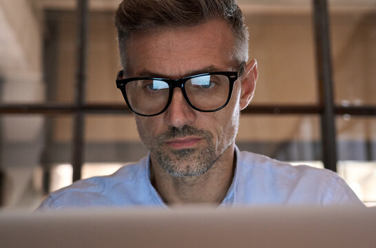 Mature concentrated business man trader wearing eyeglasses working looking at laptop computer screen reflecting in glasses analyzing online trading market financial digital data graph. Close up view