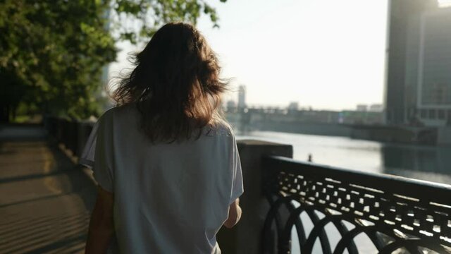 a brunette in casual clothes goes and touches hair in a city park near an openwork lattice. view from the back. the camera is moving