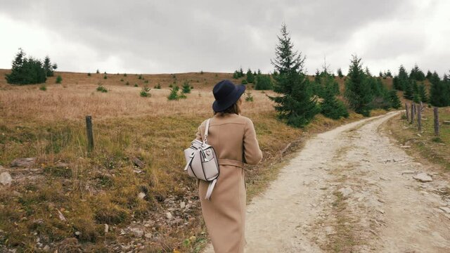 Young beautiful girl in a coat and hat walks on a field road up the hill. Tourists in mountains. Stylish cute girl carries backpack. Mountain autumn landscape. Cool cloudy weather. Rest in fresh air