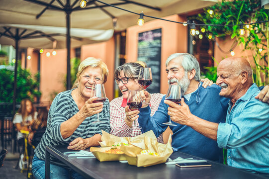 Happy elderly friends toasting with glasses of red wine sitting at a bar in the city streets - Mature people having fun celebrating in the time of happy hour - Food, drink, retirees concept