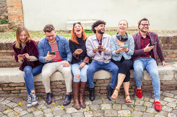 Obraz premium Group of multiracial friends sitting on a bench in the city laughing amused looking at the smart phone - Teenagers having fun and watching things on the phones - Youth addiction to technology concept