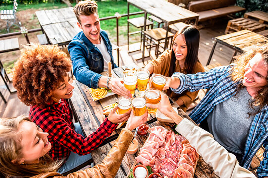 Happy friends cheering with draft beer glasses sitting around cafe bar table - Attractive young People toasting with a delicious pale ale on the terrace of the restaurant - Food and beverage concept