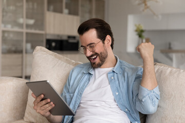 Overjoyed handsome millennial man in eyewear looking at touchpad gadget screen, celebrating online...