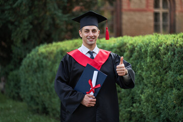 Smiling young man graduate shows like sign in graduation robe in campus with diploma